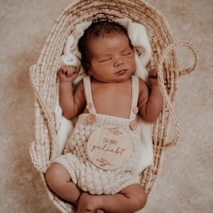 Könnte beinhalten: Ein neugeborenes Baby trägt einen cremefarbenen Strick-Strampler mit einem Holzschild, auf dem "Du bist geliebt" steht. Das Baby liegt in einem geflochtenen Korb.