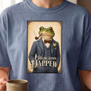 May include: Blue t-shirt featuring a framed illustration of a frog in a suit, smoking a pipe. The illustration includes the text "Feeling Kinda Dapper". The frog is wearing a blue suit, bow tie, and a flower.