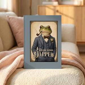 May include: A hardcover book with a light blue cover. The book's cover features a frog in a suit, smoking a pipe, with the words "Feeling Kinda Dapper". The frog is wearing a bow tie and a flower on its lapel.