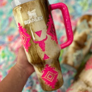 Puede incluir: Un vaso Stanley con un dise&ntilde;o de piel de vaca marr&oacute;n y crema, acentuado con dise&ntilde;os geom&eacute;tricos rosa brillante. El vaso tiene un asa rosa y una tapa blanca con pajita. El logotipo de Stanley es visible.