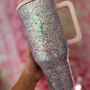 Puede incluir: Un vaso cubierto de purpurina con un asa y tapa de color rosa claro. El vaso est&aacute; cubierto de purpurina iridiscente, que refleja varios colores. El asa y la tapa son de un rosa suave y liso. El vaso est&aacute; dise&ntilde;ado para contener bebidas.