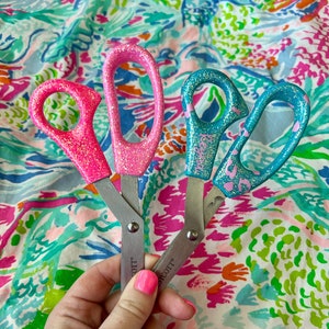 May include: Three pairs of scissors with glitter handles in pink and teal. The scissors have silver blades and are held against a colorful floral background. The handles are decorated with glitter.