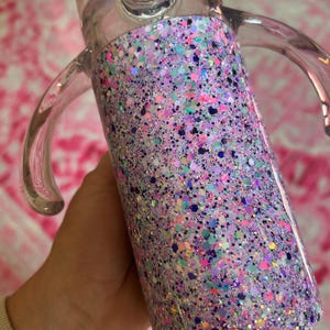 Puede incluir: Un vaso para sorber de pl&aacute;stico transparente con un dise&ntilde;o de purpurina colorido. La purpurina es una mezcla de rosa, morado, azul y verde. El vaso tiene dos asas y una tapa transparente con boquilla.