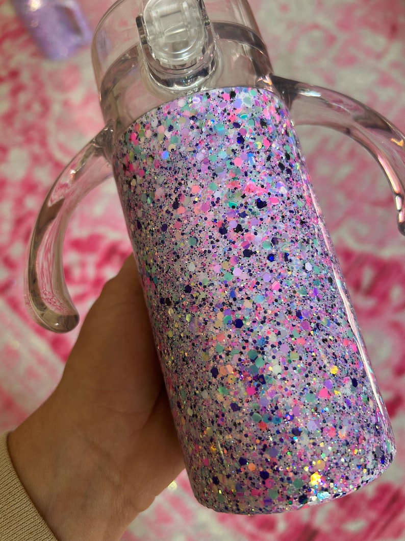 Puede incluir: Un vaso para sorber de pl&aacute;stico transparente con un dise&ntilde;o de purpurina colorido. La purpurina es una mezcla de rosa, morado, azul y verde. El vaso tiene dos asas y una tapa transparente con boquilla.