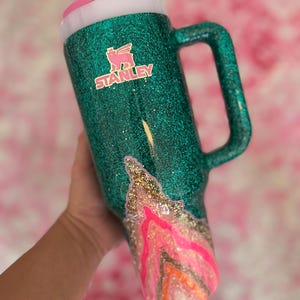 May include: A teal glitter Stanley mug with a pink and gold mountain design. The mug has a handle and a pink lid with a straw. The Stanley logo is visible on the front.