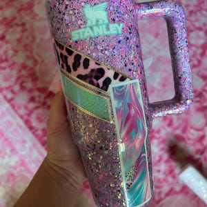 Puede incluir: Un vaso Stanley cubierto de purpurina en tonos rosa, morado y plateado. El vaso tiene un asa y un panel decorativo con estampado de leopardo, turquesa y dise&ntilde;o de arte abstracto. El logotipo de Stanley es visible.