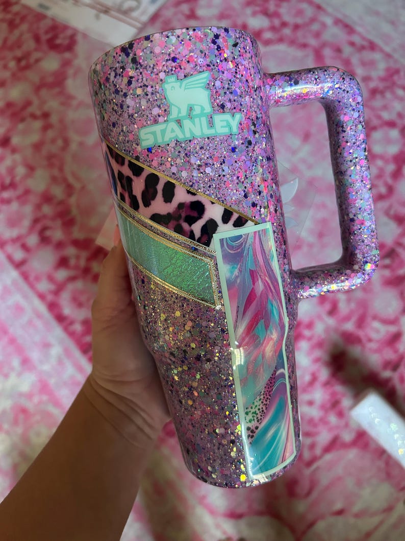 Puede incluir: Un vaso Stanley cubierto de purpurina en tonos rosa, morado y plateado. El vaso tiene un asa y un panel decorativo con estampado de leopardo, turquesa y dise&ntilde;o de arte abstracto. El logotipo de Stanley es visible.