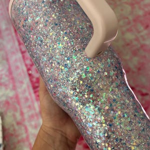 Puede incluir: Un vaso cubierto de purpurina con asa y borde rosa claro. El vaso est&aacute; cubierto con una mezcla de purpurina iridiscente y de colores pastel, creando un efecto brillante. El asa es de color rosa p&aacute;lido.