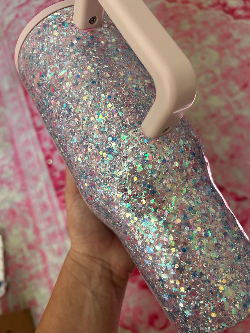 Puede incluir: Un vaso cubierto de purpurina con asa y borde rosa claro. El vaso est&aacute; cubierto con una mezcla de purpurina iridiscente y de colores pastel, creando un efecto brillante. El asa es de color rosa p&aacute;lido.