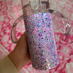 Puede incluir: Una taza para sorber de pl&aacute;stico transparente con un cuerpo cubierto de purpurina. La taza tiene una tapa transparente con cierre abatible y dos asas transparentes. El dise&ntilde;o de purpurina incluye tonos de morado, rosa, azul y verde.