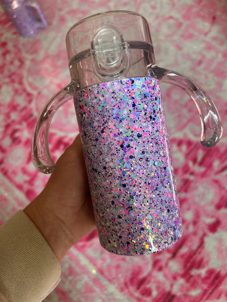 Puede incluir: Una taza para sorber de pl&aacute;stico transparente con un cuerpo cubierto de purpurina. La taza tiene una tapa transparente con cierre abatible y dos asas transparentes. El dise&ntilde;o de purpurina incluye tonos de morado, rosa, azul y verde.