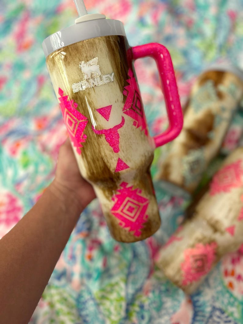 Puede incluir: Un vaso Stanley con un dise&ntilde;o de piel de vaca marr&oacute;n y crema, acentuado con dise&ntilde;os geom&eacute;tricos rosa brillante. El vaso tiene un asa rosa y una tapa blanca con pajita. El logotipo de Stanley es visible.