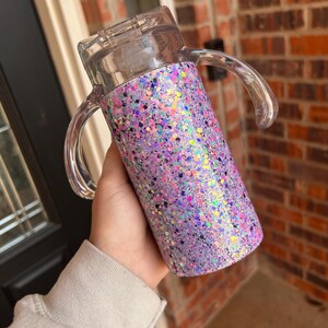 Puede incluir: Un vaso cubierto de purpurina con tapa y asas de pl&aacute;stico transparente. El vaso est&aacute; cubierto con una mezcla de purpurina morada, rosa e iridiscente. Las asas transparentes son curvas.