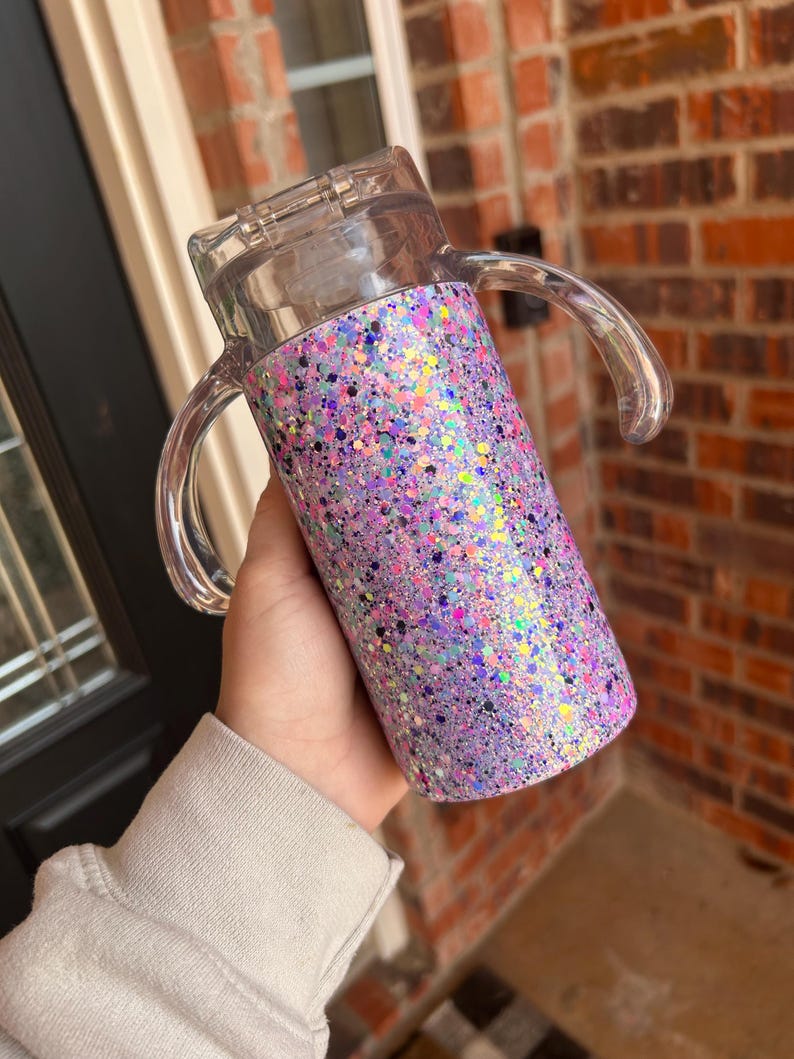 Puede incluir: Un vaso cubierto de purpurina con tapa y asas de pl&aacute;stico transparente. El vaso est&aacute; cubierto con una mezcla de purpurina morada, rosa e iridiscente. Las asas transparentes son curvas.