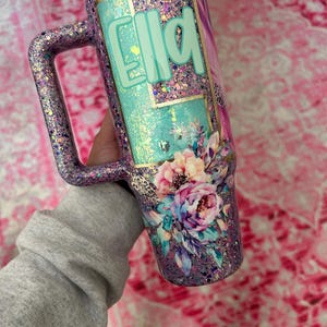 Puede incluir: Un vaso cubierto de purpurina con asa, con el nombre "Ella" en letras turquesas. El dise&ntilde;o incluye detalles florales y de estampado de leopardo, con una tapa blanca. El vaso se sostiene sobre un fondo rosa.
