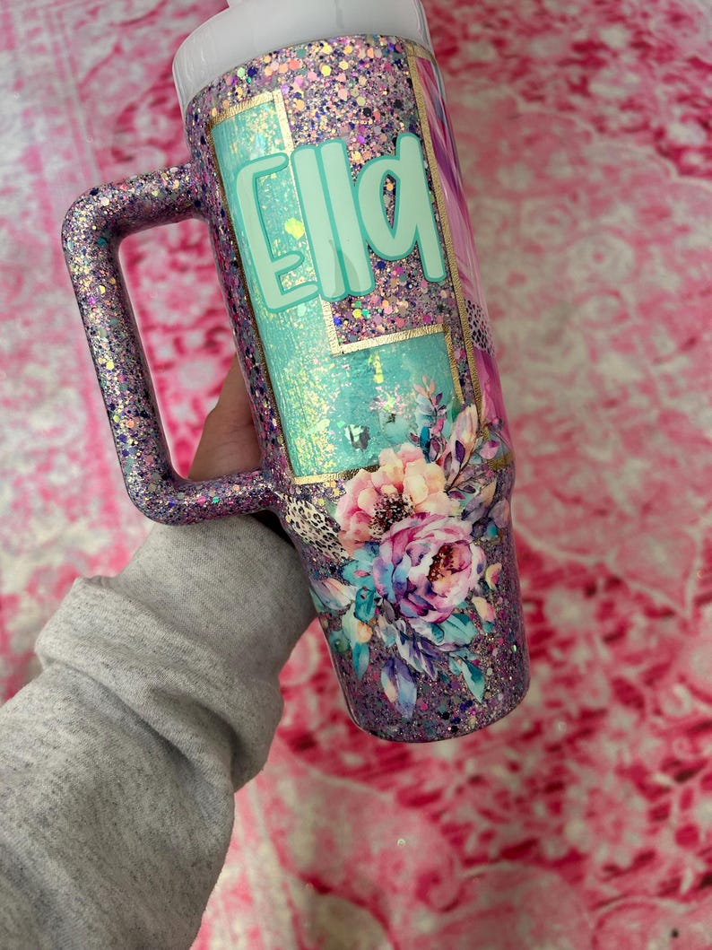 Puede incluir: Un vaso cubierto de purpurina con asa, con el nombre "Ella" en letras turquesas. El dise&ntilde;o incluye detalles florales y de estampado de leopardo, con una tapa blanca. El vaso se sostiene sobre un fondo rosa.