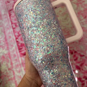 Puede incluir: Un vaso cubierto de purpurina con un asa y tapa de color rosa claro. El vaso est&aacute; cubierto de purpurina iridiscente, que refleja varios colores. El asa y la tapa son de un rosa suave y liso. El vaso est&aacute; dise&ntilde;ado para contener bebidas.