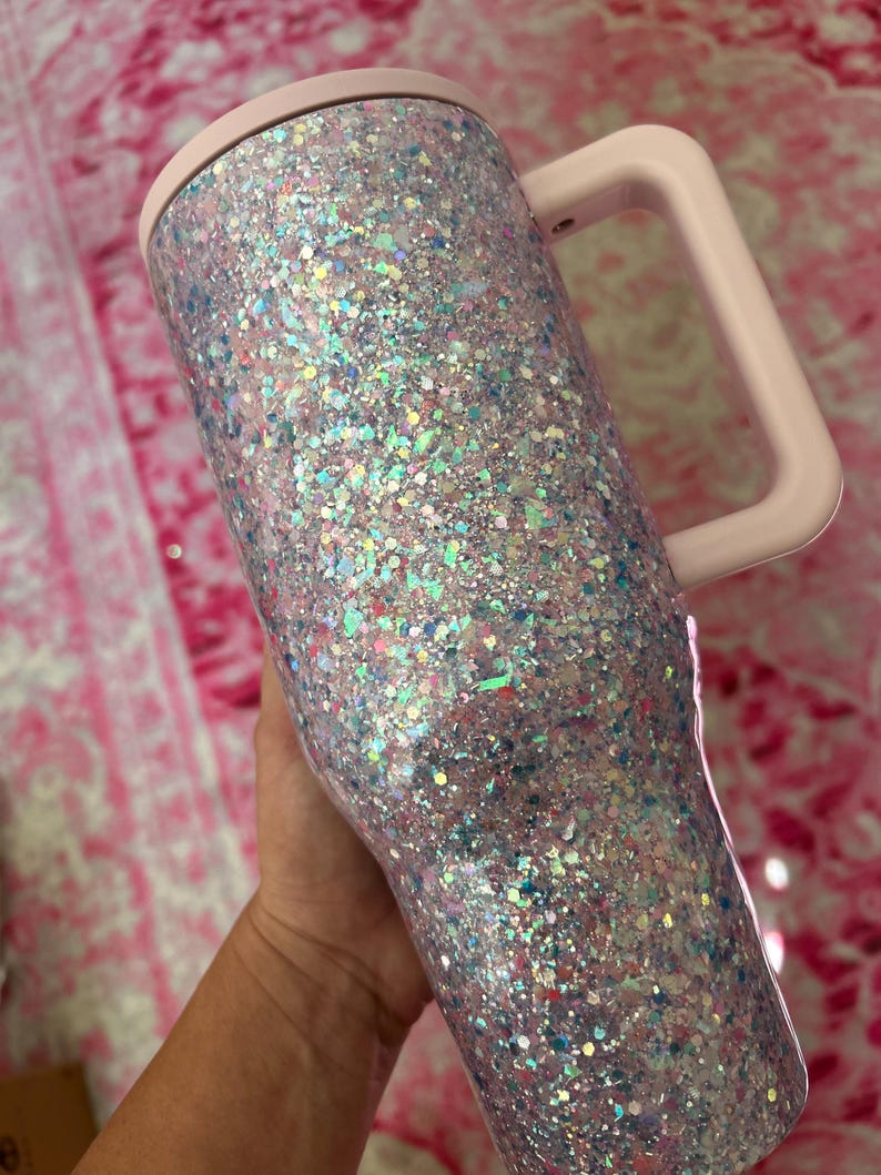 Puede incluir: Un vaso cubierto de purpurina con un asa y tapa de color rosa claro. El vaso est&aacute; cubierto de purpurina iridiscente, que refleja varios colores. El asa y la tapa son de un rosa suave y liso. El vaso est&aacute; dise&ntilde;ado para contener bebidas.