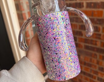 May include: A clear sippy cup with a glittery, purple, pink, and blue design. The cup has a clear lid with a flip-top closure and two clear handles. The cup is designed for toddlers and young children.