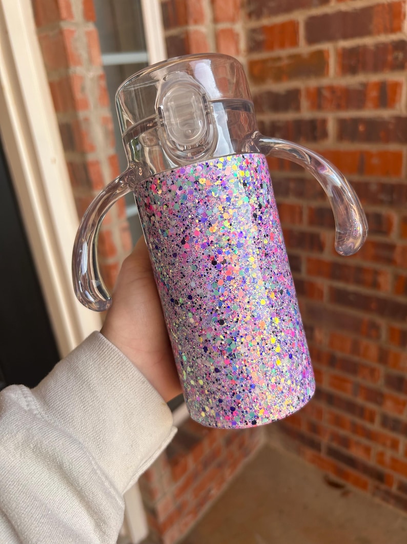 Puede incluir: Un vaso para sorber transparente con un dise&ntilde;o brillante en morado, rosa y azul. El vaso tiene una tapa transparente con cierre abatible y dos asas transparentes. El vaso est&aacute; dise&ntilde;ado para ni&ntilde;os peque&ntilde;os y ni&ntilde;os peque&ntilde;os.