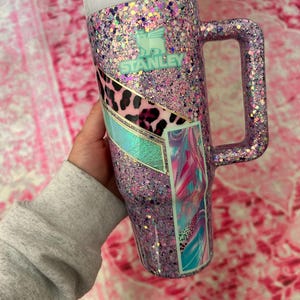 Puede incluir: Un vaso Stanley grande, cubierto de purpurina en tonos rosa y morado. El vaso tiene un asa y una tapa blanca. Los detalles incluyen un dise&ntilde;o de estampado de leopardo y el logotipo de Stanley en color turquesa. Capacidad de aproximadamente 1,18 litros.