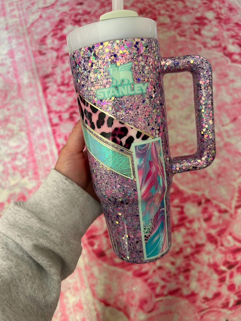 Puede incluir: Un vaso Stanley grande, cubierto de purpurina en tonos rosa y morado. El vaso tiene un asa y una tapa blanca. Los detalles incluyen un dise&ntilde;o de estampado de leopardo y el logotipo de Stanley en color turquesa. Capacidad de aproximadamente 1,18 litros.