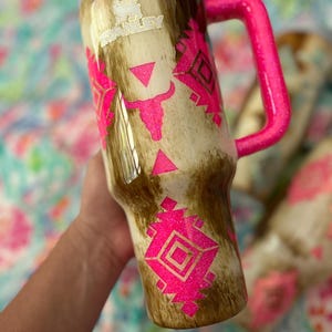 Puede incluir: Un vaso Stanley con un patr&oacute;n marr&oacute;n y beige, un asa rosa brillante y dise&ntilde;os geom&eacute;tricos rosas a juego. El vaso tiene una tapa blanca y el logotipo de Stanley. El dise&ntilde;o incluye una cabeza de toro y formas geom&eacute;tricas.