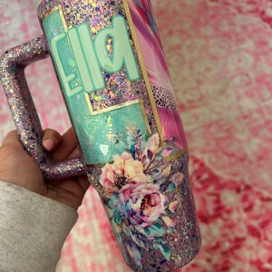 Puede incluir: Vaso cubierto de purpurina con asa, con el nombre "Ella" en verde menta. El dise&ntilde;o incluye detalles florales y una mezcla de purpurina rosa, morada y dorada. El vaso tiene una tapa blanca y un asa cubierta de purpurina.