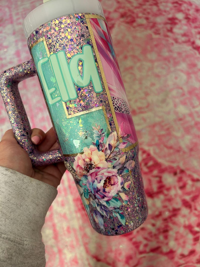 Puede incluir: Vaso cubierto de purpurina con asa, con el nombre "Ella" en verde menta. El dise&ntilde;o incluye detalles florales y una mezcla de purpurina rosa, morada y dorada. El vaso tiene una tapa blanca y un asa cubierta de purpurina.