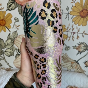 May include: A pink glitter tumbler with a tropical and leopard print design. The tumbler features gold and green leaves, brown leopard spots, and a pink glitter background.