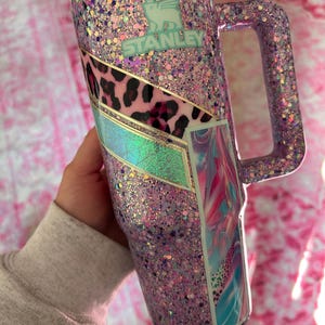 Puede incluir: Un vaso Stanley cubierto de purpurina en tonos rosa, morado y purpurina iridiscente. El vaso presenta un asa y un panel decorativo con estampado de leopardo y un dise&ntilde;o abstracto colorido. El logotipo de Stanley es visible.