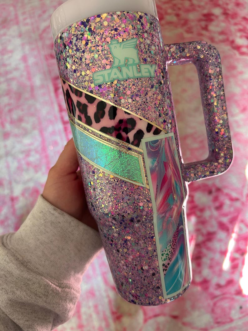 Puede incluir: Un vaso Stanley cubierto de purpurina en tonos rosa, morado y purpurina iridiscente. El vaso presenta un asa y un panel decorativo con estampado de leopardo y un dise&ntilde;o abstracto colorido. El logotipo de Stanley es visible.
