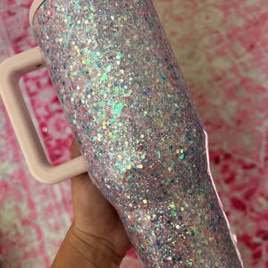 Puede incluir: Un vaso cubierto de purpurina con asa y tapa rosa claro. El vaso est&aacute; cubierto de purpurina iridiscente que refleja m&uacute;ltiples colores. El asa y la tapa son de color rosa suave liso. El vaso est&aacute; dise&ntilde;ado para contener bebidas.