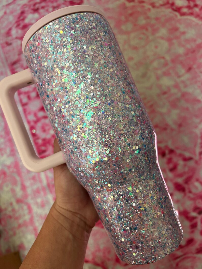 Puede incluir: Un vaso cubierto de purpurina con asa y tapa rosa claro. El vaso est&aacute; cubierto de purpurina iridiscente que refleja m&uacute;ltiples colores. El asa y la tapa son de color rosa suave liso. El vaso est&aacute; dise&ntilde;ado para contener bebidas.