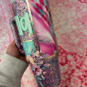 Puede incluir: Un vaso con purpurina y asa, con un dise&ntilde;o floral y la palabra "wild" en verde menta. El vaso tiene una combinaci&oacute;n de colores rosa, azul y dorado, con una tapa blanca y una pajita.