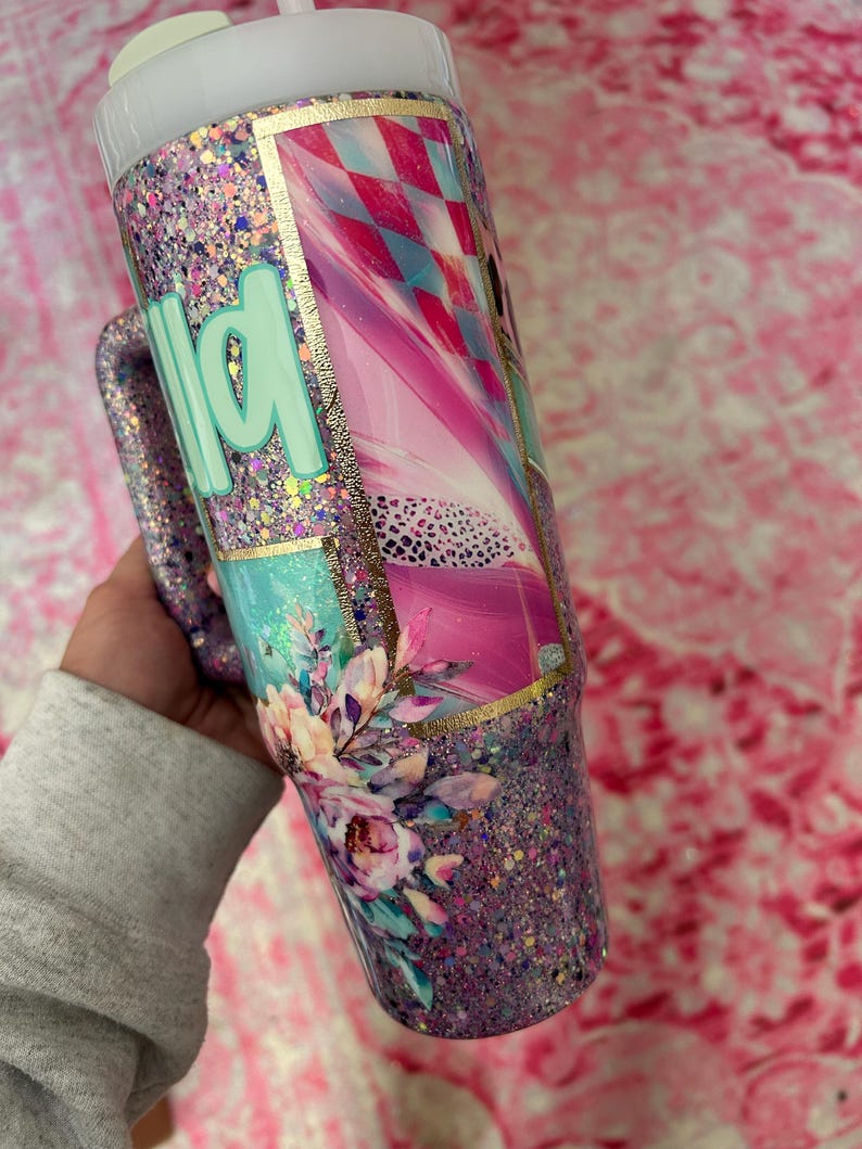 Puede incluir: Un vaso con purpurina y asa, con un dise&ntilde;o floral y la palabra "wild" en verde menta. El vaso tiene una combinaci&oacute;n de colores rosa, azul y dorado, con una tapa blanca y una pajita.