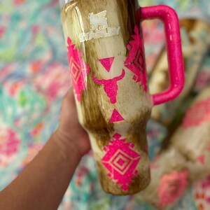 Puede incluir: Un vaso grande y aislado con tapa blanca y asa rosa brillante. El vaso presenta un patr&oacute;n marr&oacute;n y beige con dise&ntilde;os geom&eacute;tricos rosas y una calavera de toro. El logotipo de Stanley es visible.