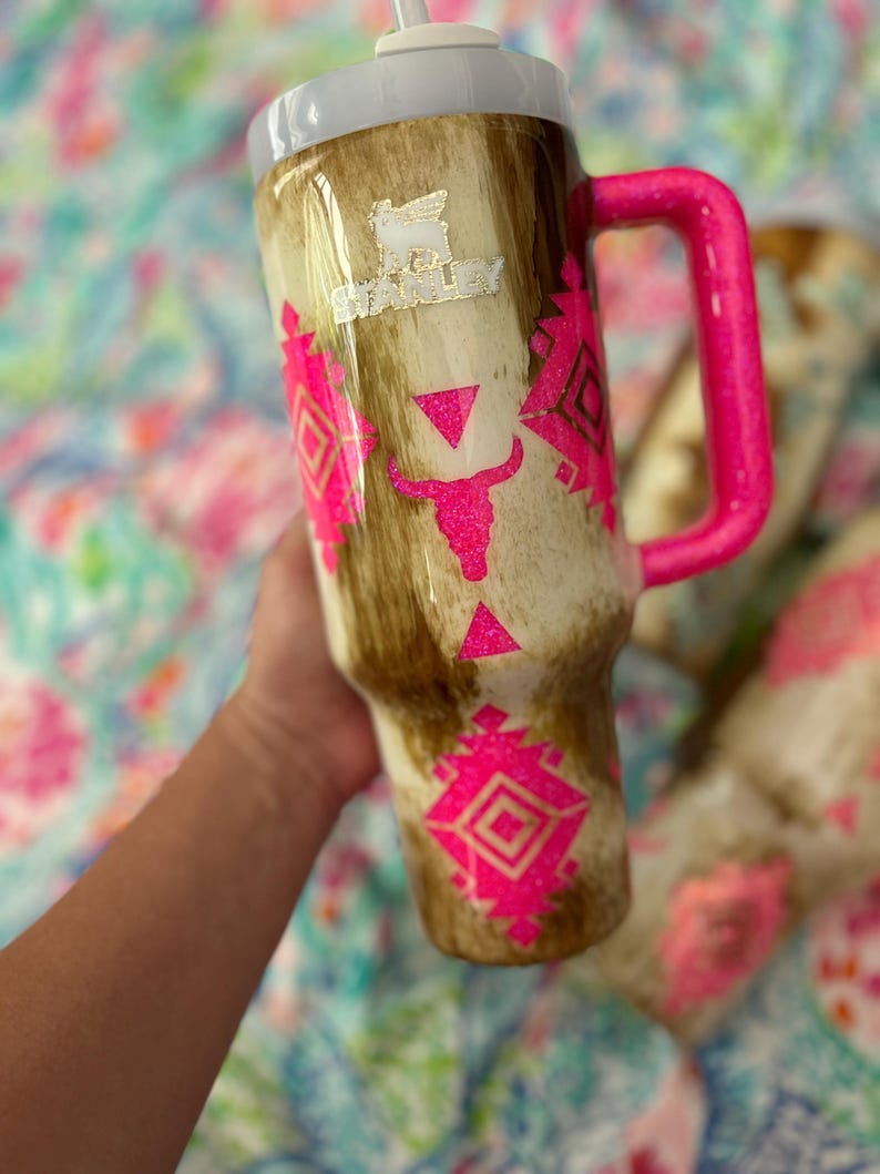 Puede incluir: Un vaso grande y aislado con tapa blanca y asa rosa brillante. El vaso presenta un patr&oacute;n marr&oacute;n y beige con dise&ntilde;os geom&eacute;tricos rosas y una calavera de toro. El logotipo de Stanley es visible.