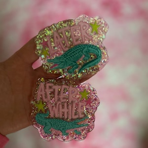 May include: Two glittery resin alligator pins. One pin says "Later" with a teal alligator and pink text. The other pin says "After While" with a teal alligator and pink text. Both pins have yellow and pink star accents.
