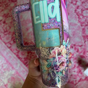 Puede incluir: Un vaso con purpurina y asa, con el nombre "Ella" en turquesa. El dise&ntilde;o incluye motivos florales, salpicaduras de morado, rosa y dorado, y un acabado texturizado y brillante. El vaso est&aacute; dise&ntilde;ado para contener una bebida.