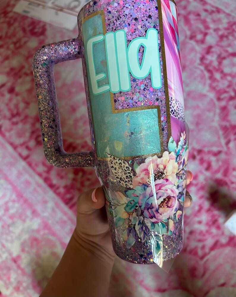 Puede incluir: Un vaso con purpurina y asa, con el nombre "Ella" en turquesa. El dise&ntilde;o incluye motivos florales, salpicaduras de morado, rosa y dorado, y un acabado texturizado y brillante. El vaso est&aacute; dise&ntilde;ado para contener una bebida.