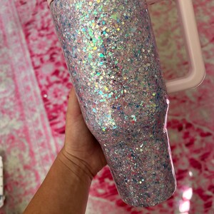 Puede incluir: Un vaso cubierto de purpurina con asa, tapa y borde de color rosa claro. El vaso est&aacute; cubierto de purpurina iridiscente en tonos rosa, azul y plata. El asa es cuadrada y la tapa es plana.