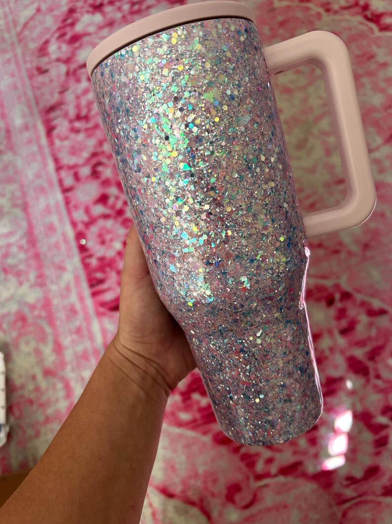 Puede incluir: Un vaso cubierto de purpurina con asa, tapa y borde de color rosa claro. El vaso est&aacute; cubierto de purpurina iridiscente en tonos rosa, azul y plata. El asa es cuadrada y la tapa es plana.