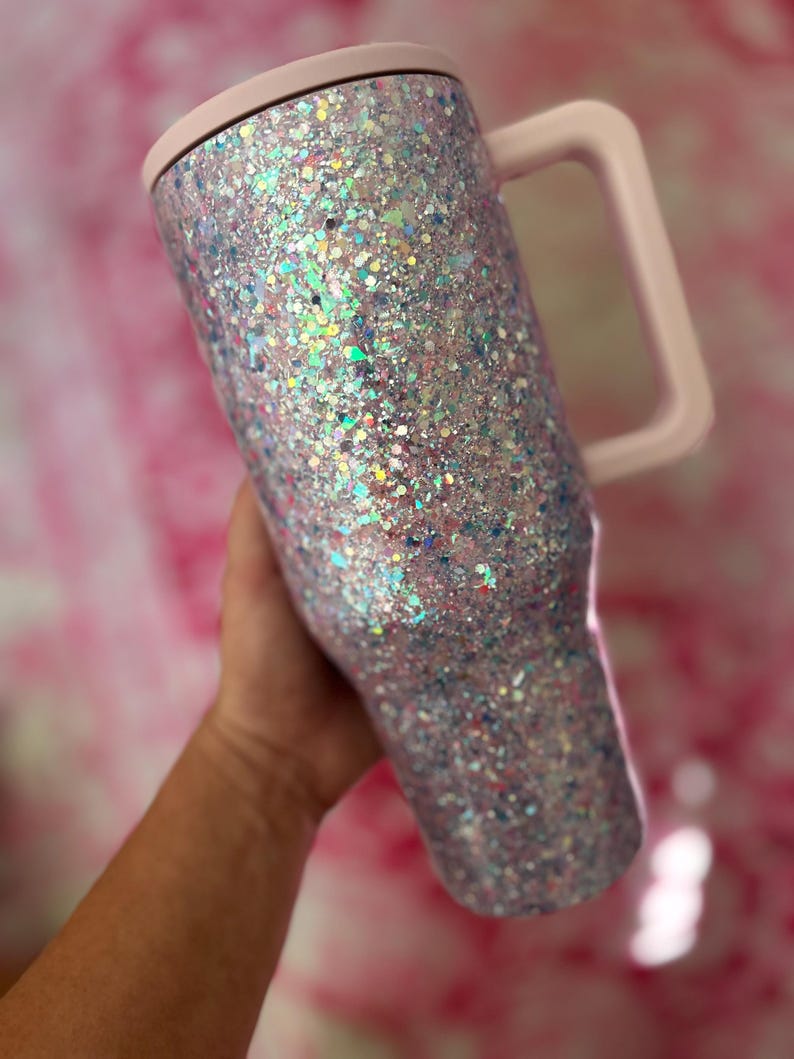 Puede incluir: Un vaso cubierto de purpurina con un asa y tapa de color rosa claro. El vaso est&aacute; cubierto de purpurina iridiscente, que refleja varios colores. El asa y la tapa son de un rosa suave y liso. El vaso est&aacute; dise&ntilde;ado para contener bebidas.