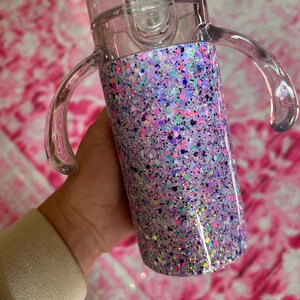 Puede incluir: Una taza para sorber transparente con una base cubierta de purpurina. La base est&aacute; cubierta con una mezcla de purpurina rosa, morada, azul y plateada. La taza tiene dos asas transparentes y una tapa transparente con cierre abatible.