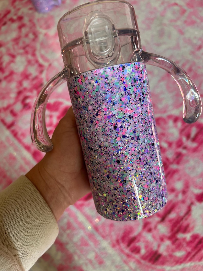 Puede incluir: Una taza para sorber transparente con una base cubierta de purpurina. La base est&aacute; cubierta con una mezcla de purpurina rosa, morada, azul y plateada. La taza tiene dos asas transparentes y una tapa transparente con cierre abatible.