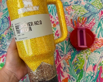 May include: A yellow glitter tumbler with a black and silver glitter design. The tumbler has a handle and a label that reads "The Flow State Quencher H2.0 Tumbler 40 oz | 1.18 L All Day Hydration".