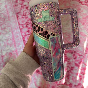 Puede incluir: Un vaso Stanley cubierto de purpurina en tonos morados, rosas y acentos iridiscentes. El vaso tiene un asa, una tapa y una pajita. El logotipo de Stanley es visible. Paneles decorativos con estampado animal y dise&ntilde;os abstractos a&ntilde;aden inter&eacute;s visual.