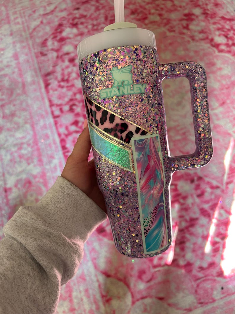 Puede incluir: Un vaso Stanley cubierto de purpurina en tonos morados, rosas y acentos iridiscentes. El vaso tiene un asa, una tapa y una pajita. El logotipo de Stanley es visible. Paneles decorativos con estampado animal y dise&ntilde;os abstractos a&ntilde;aden inter&eacute;s visual.