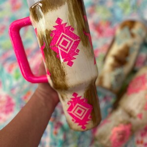 Puede incluir: Un vaso con un patr&oacute;n de piel de vaca marr&oacute;n y blanco y un asa rosa brillante. El vaso presenta un dise&ntilde;o geom&eacute;trico rosa. La tapa es blanca y se ve una pajita blanca. El vaso est&aacute; dise&ntilde;ado para contener una bebida.